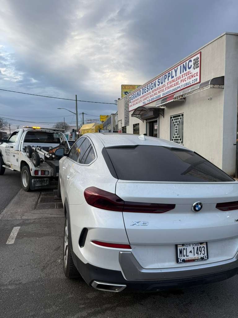 BMW X6 flatbed run past a Queens commercial strip White BMW X6 being towed past Grand Design Supply on a Queens commercial street — JG Towing.